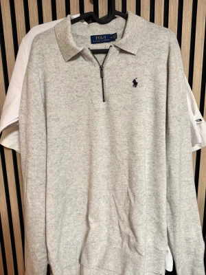 Ljusgrå stickad tröja Polo Ralph Lauren - Snygg ljusgrå stickad tröja från Polo Ralph Lauren med halv dragkedja och klassisk krage. Tröjan har en broderad marinblå logga på bröstet och ribbade muddar. Perfekt för en clean och stilren look.