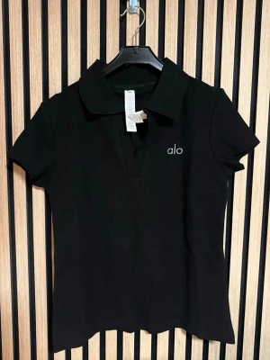 Svart polotopp från Alo - Svart polotopp från Alo med klassisk krage och korta ärmar. Diskret Alo-logga broderad på bröstet. Toppens material är mjuk bomull och passformen är normal. Perfekt för dig som gillar stilrena och enkla plagg.