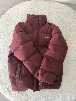 Vinröd pufferjacka från Carhartt - Snygg vinröd pufferjacka från Carhartt med hög krage och diskret logga på bröstet som knappt är använd. Jackan har två sidofickor, dragkedja och tryckknappar framtill samt elastiska muddar vid ärmsluten. Perfekt för kalla dagar och riktigt trendig look. Nypris 3800