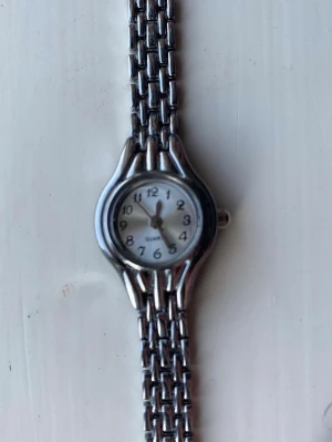 Silverfärgad klassisk armbandsklocka - Stilren armbandsklocka i silverfärg med rund urtavla och tydliga svarta siffror. Klockan har en länkad metallrem och enkel, klassisk design som passar till det mesta. Urtavlan är vit och klockan drivs av quartzverk.