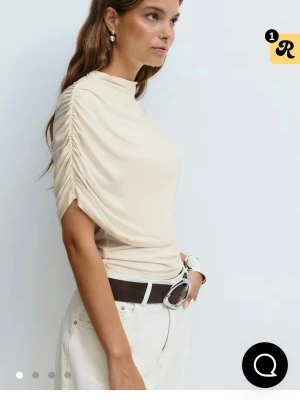 Beige omlottopp med rynkade ärmar - Trendig creme vit poncho topp med rynkade korta ärmar. Perfekt för dig som gillar stilrena och moderna plagg. Toppen har en hög hals och är gjord i ett mjukt material som sitter snyggt på kroppen.