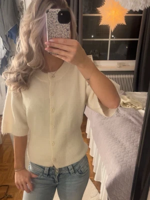 Krämvit kofta med kort ärm - Söt krämvit kofta med korta ärmar och rund halsringning. Koftan har knappar framtill och är stickad i ett mjukt material som ger en mysig känsla. Perfekt att styla med jeans eller kjol för en enkel men chic look.