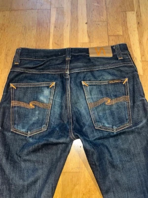 Mörkblå jeans från Nudie Jeans - Säljer ett par mörkblå jeans från Nudie Jeans med klassiska kontrastsömmar och vågdetaljer på bakfickorna. Jeansen har en smal passform och är tillverkade i slitstark denim. Perfekta för dig som gillar en tidlös och snygg look.