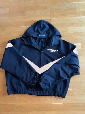 Svart Essentials tröja med vit detalj - Snygg svart hoodie från Essentials med dragkedja och vit V-formad detalj över bröstet och armarna. Huva och elastisk midja ger en sportig vibe. Perfekt för dig som gillar streetwear och enkel stil. Det enda nackdelen är den lilla fläcken på tröjan. 