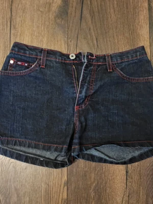 Mörkblå jeansshorts från C'est Toi - Snygga mörkblå jeansshorts från C'est Toi med röda kontrastsömmar och klassisk femficksdesign. Shortsen har normal passform, midwaist och stängs med dragkedja och knapp. Perfekta för sommaren och lätta att matcha med olika stilar.