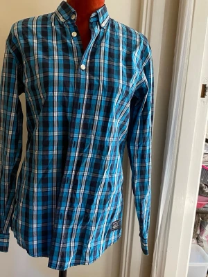 Blå- och svart-rutig skjorta S - Snygg långärmad skjorta med blå, svart och vit rutigt mönster. Skjortan har button-down krage och vita knappar framtill. Perfekt för dig som gillar en klassisk men ändå färgstark stil. Materialet känns lätt och bekvämt.
