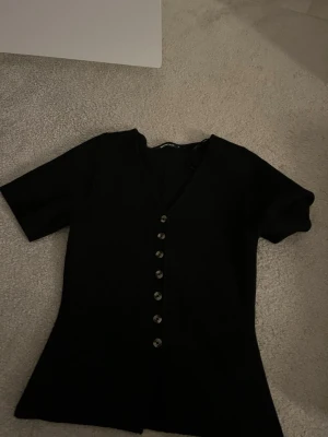 Svart v-ringad topp med knappar - Snygg  stickad svart topp med v-ringning och dekorativa knappar framtill. Kortärmad modell i mjukt material som sitter bekvämt och har en stilren look. Perfekt att matcha med jeans eller kjol för en trendig outfit.