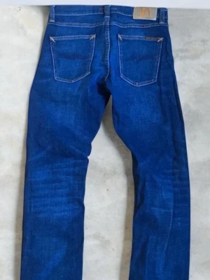 Nudie Jeans - Snygga blå jeans från Nudie Jeans med klassisk femficksmodell och raka ben. Jeansen har kontrastsömmar och en patch bak i midjan. Tillverkade i slitstark denim som ger en cool och tidlös look. Endast använda ett fåtal gånger. Priset är ej hugget i sten. W:27 L:32