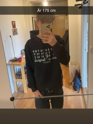 Mörkblå hoodie från Maison Margiela - Maison Margiela hoodie i mörkblå med vit grafisk print på framsidan. Klassisk huva med dragsko och ribbade muddar. Materialet är mjukt och bekvämt, perfekt för en avslappnad look. Paris-logga under printen ger extra stilpoäng.