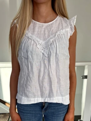 Topp med spets - Säljer en söt vit blus från Zara med spetsdetaljer framtill och volang vid ärmslut. Blusen är ärmlös och har en liten knapp i nacken. Storlek S från Zara.