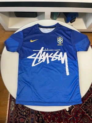 Blå Stüssy x Brasil t-shirt - Säljer en blå t-shirt i samarbete mellan Stüssy och Brasil, med stor vit Stüssy-logga framtill och Nike-märke. Baksidan har en färgstark grafik med två tecknade figurer och texten 'WAiROON DURIAN'. Perfekt för dig som gillar streetwear och unika prints.
