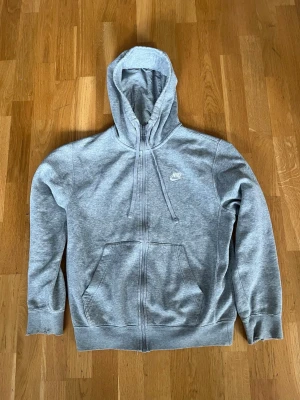 Grå zip hoodie från Nike - Klassisk grå zip hoodie från Nike med huva och dragsko. Hoodien har fickor framtill och Nike-logga broderad på bröstet. Tillverkad i mjuk bomullsblandning, perfekt för chill dagar eller till träningen. Storlek xs men passar till S också