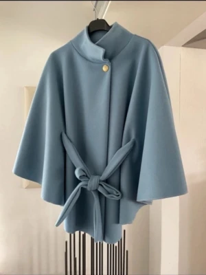 Ljusblå cape-kappa med knytband - Stilren ljusblå cape-kappa med knytband i midjan och en dekorativ guldknapp vid halsen. Kappan har vid passform, korta vida ärmar och hög krage. Perfekt för dig som vill ha en trendig och elegant look. Materialet ser ut att vara mjukt och tjockt, troligen ullblandning.