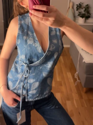 Blå omlottopp med blommigt mönster - Säljer en blå omlottopp i denim-look med vitt blommigt mönster. Toppen är ärmlös, har djup v-ringning och knyt i sidan för en snygg passform. Perfekt för dig som vill ha en unik och trendig topp till jeans eller kjol.