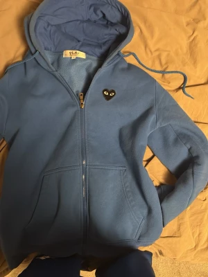 Blå hoodie från Comme des Garçons Play - Säljer en blå hoodie från Comme des Garçons Play med dragkedja, huva och snörning. På bröstet finns det klassiska svarta hjärtat med ögon och en större version av hjärtat på ryggen. Mjuk bomullskvalitet och två fickor framtill.