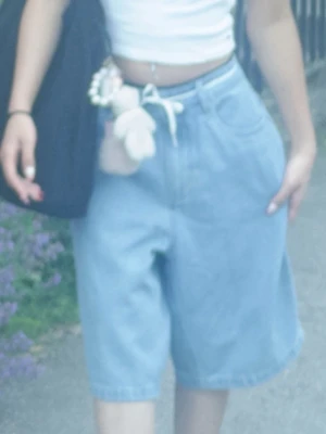 Vida ljusblå jeansshorts med snörning - Säljer ett par vida ljusblå jeansshorts med hög midja och vit snörning i midjan. Shortsen har klassisk femficksdesign och är tillverkade i denim. Perfekta för en avslappnad och trendig stil.