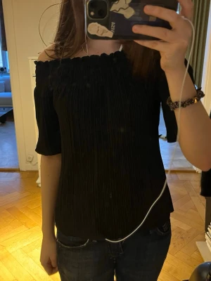 Svart offshoulder topp med plissering - Säljer en svart offshoulder topp med plisserad struktur och korta ärmar. Toppen har en elastisk kant upptill som ger en snygg passform och trendig look. Perfekt för dig som vill ha en stilren och modern topp till garderoben.
