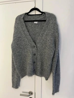 Grå fluffig kofta med pärlor - Mysig grå kofta med fluffig känsla och dekorativa pärlor längs axlar och framkant. Koftan har v-ringning, knappar framtill och långa ärmar. Perfekt till våren och sena sommarkvällar 