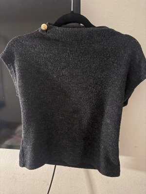 Mörkgrå stickad topp med knappdetalj - Säljer en mörkgrå stickad topp med korta ärmar och en dekorativ träknapp vid axeln. Toppen har en rak och boxig passform och är tillverkad i ett mjukt, stickat material. Perfekt för lager-på-lager och enkel att styla till olika outfits.Från SHEIN i storlek S