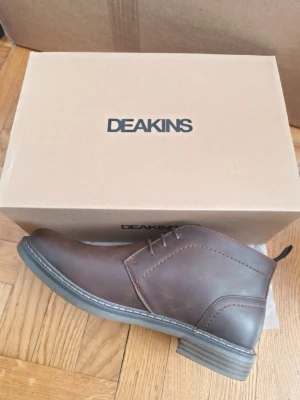 Bruna läderstövletter från Deakins - Snygga bruna stövletter från Deakins i slätt läder med diskreta sömmar och klassisk snörning. Skorna har en robust sula och en bekväm, vadderad insida med rutigt foder. Perfekta för dig som vill ha stilrena boots med modern touch.