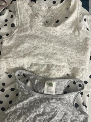 2 spetsbhar/toppar, 134/140 - Två toppar från Lindex: en vit linnetopp i spets med blommigt mönster och racerback, samt en grå linnetopp med vita hjärtan och sportig rygg. Båda är ärmlösa och har en croppad passform, perfekta för att ge en chill vibe till din outfit. 💕Använda fåtal gånger! inga defekter!💕båda är 134/140 även om de bara är bild på enas storlek. Vad jag vet så är den gråa från Lindex! 