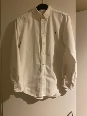 Vit skjorta från Ralph Lauren - Barnstorlek 14 custom fit. Inga konstigheter. Det är en klassisk vit skjorta från Ralph Lauren med button-down krage och broderad logga på bröstet. Skjortan har långa ärmar, knappar framtill och är tillverkad i mjuk bomull. Perfekt för en clean och stilren look. Skriv vid frågor!