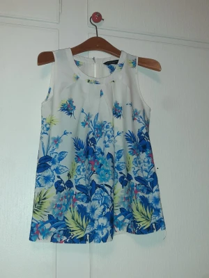 Blommig ärmlös topp i blått och vitt från Dorothy Perkins  - Snygg ärmlös topp med blommigt mönster i blått, grönt och vitt. Toppen har rund halsringning och lätt utsvängd passform. Perfekt för varma dagar och ger en fräsch sommarkänsla. Tillverkad i ett lätt och luftigt material.