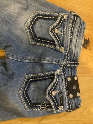 Ljusblå Miss Me jeans med detaljer - Säljer ett par ljusblå Miss Me jeans med slitningar och snygga kontrastsömmar. Bakfickorna har dekorativa broderier, nitar och blingdetaljer. Jeansen har bootcut passform och är perfekta för dig som gillar statement-plagg. Köpta här på pick men tyvärr för små. Mina dröm jeansss. Passar någon mindre!