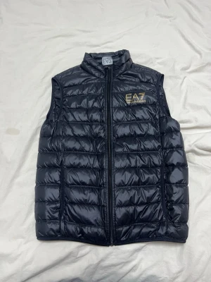 Svart dunväst från EA7 Emporio Armani - Snygg svart dunväst från EA7 Emporio Armani med guldfärgad logga på bröstet. Västen har hög krage, dragkedja framtill och quiltad design som ger en sportig look. Perfekt att slänga över en hoodie eller tröja för extra värme.