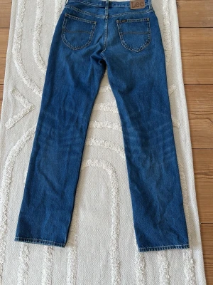 Blå raka jeans från Lee W30 L32 - Snygga blå jeans från Lee med raka ben och klassiska bakfickor. Jeansen har en enkel design med Lee-logga på bakfickan och är tillverkade i bomullsdenim. Perfekta för en avslappnad och stilren look.