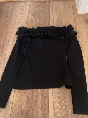 Svart offshoulder topp från NLY Trend - Trendig svart offshoulder topp från NLY Trend i stretchigt material. Toppen har långa ärmar och en bred volang längs den axelbara kanten som ger en snygg och modern look. Perfekt att styla med höga jeans eller kjol.