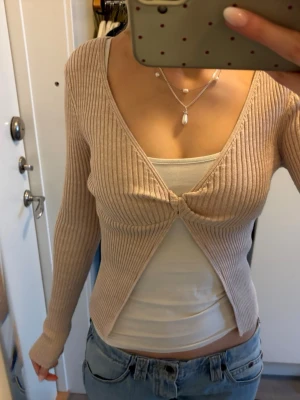 Beige ribbad omlottkofta - Trendig beige ribbad omlottkofta med lång ärm och djup v-ringning. Koftan har twistad detalj framtill och är tillverkad i ett stretchigt, mjukt material som sitter snyggt på kroppen. Perfekt att styla med linne eller topp under.