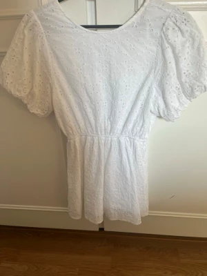 Vit kort klänning med puffärm - Söt vit kortklänning med puffärmar och broderade blommor. Klänningen har rund halsringning och markerad midja som ger en fin siluett. Tillverkad i bomull för en luftig och bekväm känsla. Perfekt för sommarens alla tillfällen!