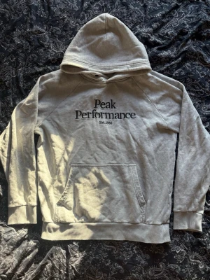 Grå hoodie från Peak Performance - Snygg grå hoodie från Peak Performance med svart logga och text 'Est. 1986' på bröstet. Klassisk känguruficka framtill, mjuk insida och ribbade muddar vid ärmar och nederkant. Perfekt för en avslappnad och sportig stil.