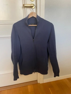 Mörkblå ziptröja från Massimo Dutti - Mörkblå ziptröja från Massimo Dutti med hög krage och två fickor framtill. Tröjan har en stilren design med ribbade muddar vid ärmslut och nederkant. Perfekt för lager på lager och enkel att matcha med olika outfits.