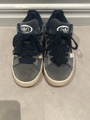 Adidas Campus mörkblå sneakers - Mörkblå Adidas Campus sneakers med klassiska vita ränder på sidan och Adidas-logga på plösen. Ovandelen är i mocka och sulan är vit med gummidetaljer. Skorna har breda mörkblå skosnören och låg profil för en clean och tidlös look.