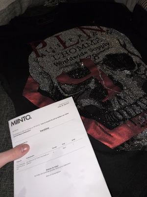 Svart t-shirt med dödskalle från Philipp Plein - Storlek S. Cool svart t-shirt från Philipp Plein med stort dödskallemotiv i silver och röda detaljer på framsidan. T-shirten har korta ärmar och är tillverkad i bomull. Perfekt för dig som vill sticka ut med en edgy look. Äkta, kvitto ses i bilden. Sparsamt använd