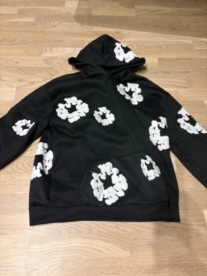 Svart hoodie med vita blommor - Snygg svart hoodie från Denim Tears med stora vita blommor tryckta över hela plagget. Klassisk huva och magficka framtill. Perfekt för dig som vill sticka ut med en unik och trendig stil. Materialet känns mjukt och bekvämt, perfekt för vardagslooken.