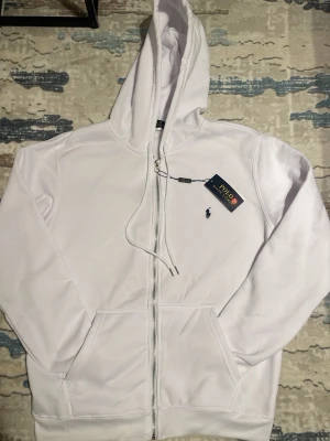 Vit hoodie från Polo Ralph Lauren - Snygg vit hoodie från Polo Ralph Lauren med dragkedja framtill och klassisk blå logga broderad på bröstet. Hoodien har huva med dragsko och två fickor framtill. Tillverkad i mjukt bomullsmaterial som känns skönt mot huden. Perfekt för en clean och stilren look.