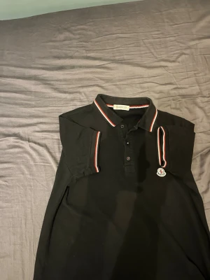 Svart Moncler pikétröja med ränder - Säljer en svart pikétröja från Moncler med klassisk krage och knappar framtill. Tröjan har röda och vita ränder på krage och ärmslut samt Moncler-logga på bröstet. Perfekt för dig som gillar stilrena och sportiga plagg. Pris kan diskuteras.