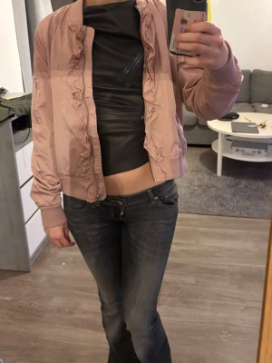 Ljusrosa bomberjacka från Magiano - Säljer en ljusrosa bomberjacka från Magiano med volangdetaljer längs dragkedjan. Jackan har ribbade muddar och är perfekt för dig som vill ha en söt och trendig look. Passar till många olika outfits och har en skön, normal passform.