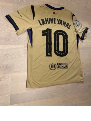 FC Barcelona Lamine Yamal matchtröja - Säljer en officiell FC Barcelona matchtröja med Lamine Yamal och nummer 10 på ryggen. Tröjan är guld på baksidan och har klassiska blå-röda ränder framtill, Champions League-märke på ärmen och Spotify-logga på bröstet. Tillverkad i lätt och ventilerande polyester. 2x
