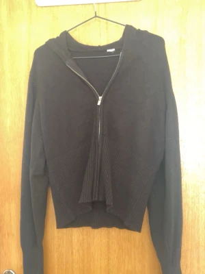 Svart stickad hoodie med dragkedja - Säljer en svart stickad hoodie med dragkedja framtill och ribbade detaljer vid midjan och ärmsluten. Hoodien har en croppad passform och långa ärmar. Perfekt för dig som gillar en enkel men snygg stil.