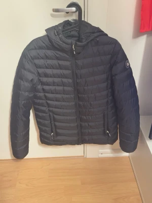 Svart pufferjacka med huva - Snygg svart pufferjacka med huva och dragkedja framtill. Jackan har två fickor med dragkedja och quiltad design. Märke syns på ärmen. Perfekt för kalla dagar och riktigt skön att bära. Jackan har små defekter, hör av dig för bilder.