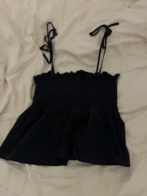  linnetopp med knytband - Söt svart linnetopp med volangkant och smockad överdel. Axelbanden är tunna och knyts upptill för en justerbar passform. Perfekt till sommaren och enkel att styla med jeans eller kjol.