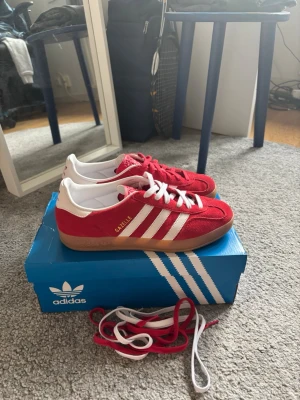 Adidas Gazelle röda sneakers - Säljer ett par klassiska Adidas Gazelle sneakers i rött mocka med vita detaljer och gummisula. Skorna har de ikoniska tre vita ränderna på sidan, guldfärgad Gazelle-text och kommer med både röda och vita skosnören. Perfekt för dig som gillar retrostil och streetwear.