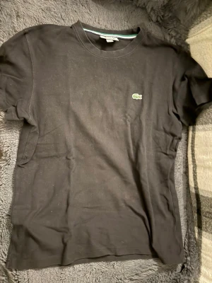 Svart t-shirt från Lacoste - Klassisk svart t-shirt från Lacoste med rund hals och den ikoniska gröna krokodilloggan broderad på bröstet. Tillverkad i mjuk bomull och har en normal passform. Perfekt basplagg för en clean och stilren look.