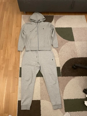 Grå mjukisdress från Polo Ralph Lauren - Säljer en grå mjukisdress från Polo Ralph Lauren med dragkedja på hoodien och matchande byxor. Setet har små broderade loggor på både tröja och byxa, ribbade muddar och justerbar huva. Perfekt för chill dagar hemma eller när du vill vara bekväm.
