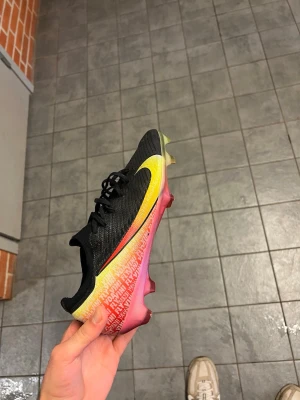 Nike Mercurial fotbollsskor svart/gul - Snygga Nike Mercurial fotbollsskor i svart med stora gula swoosh-loggor och färgskiftande detaljer i gult, grönt och rosa. Ovandelen är tillverkad i ett stickat syntetmaterial med texten 'FLYKNIT' tryckt längs sidan. Skorna är slutsålda! Vini JR Vini Fly. Ytterligare ett par limited edition skor från Vinicius JR som är i jättebra skick. Storlek 44. Säljs pågrund av att jag tappat intresset av spelaren. Jättefrsächa dubbar och använda några gånger bara. Vid intresse skickas bilder! Köpta för 3499