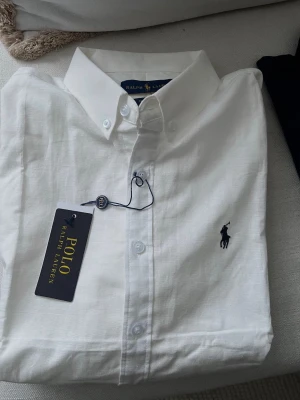Vit skjorta från Polo Ralph Lauren - Klassisk vit skjorta från Polo Ralph Lauren med button-down krage och broderad marinblå logga på bröstet. Tillverkad i tunn bomull, långärmad och med normal passform. Perfekt till jeans eller chinos för en clean och stilren look. ( den är inte äkta men scanner funkar och du får med pris lapp) Jag är 170 och väger 58